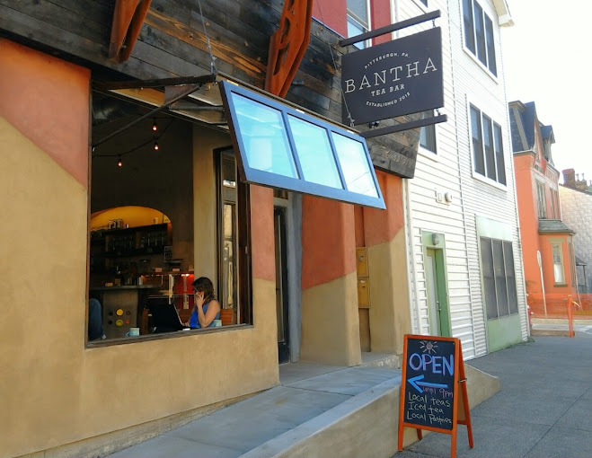 Tea shop with a rustic facade of horizontal boards over a large open window. Underneath the boards, the building is painted burnt orange and mustard yellow. There are two signs out front. One sign reads "Bantha Tea Bar," and the other sign reads "Open until 9 p.m. Local teas, Iced tea, Local pastries."