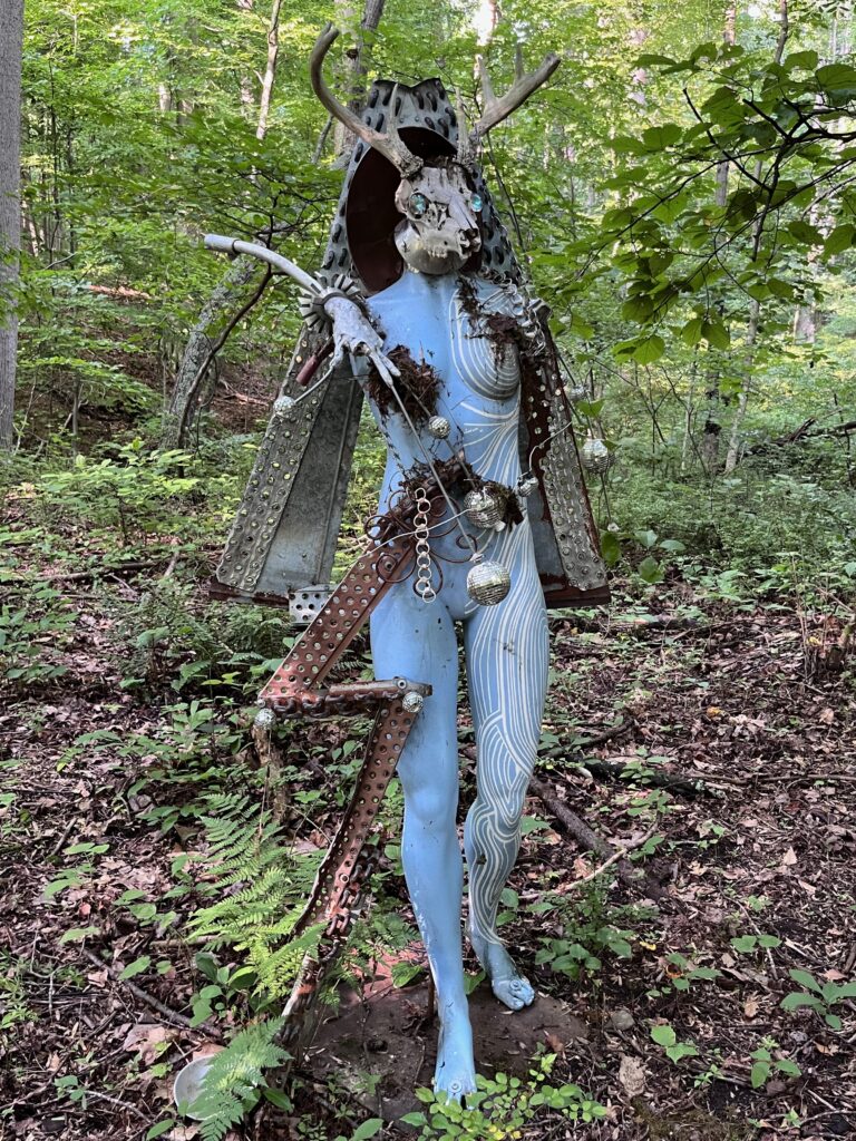 A modern art outdoor sculpture with a woman's body painted blue with white circular lines, a deer's head, a cape made out of metal, and something that looks like a bow and arrow.